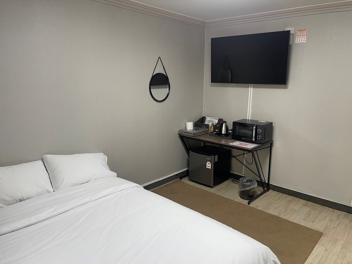 A simple sleeping area features a spacious bed with white linens, situated next to a modern television mounted on the wall. A small table hosts a kettle, microwave, and refrigerator, enhancing the functionality of the space. A round mirror adds subtle design to the room.