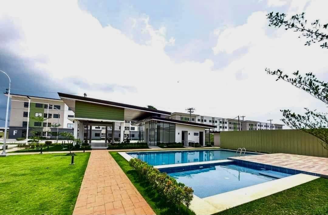 A spacious outdoor area features a well-maintained swimming pool surrounded by lush green grass. A walkway paved with terracotta tiles leads toward a modern building with large glass windows. The serene environment is complemented by gentle clouds in a clear sky.