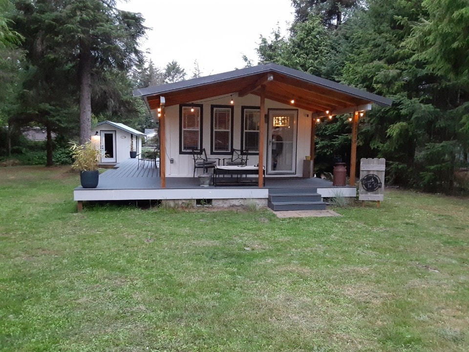 The exterior of a charming house is showcased, featuring a covered porch with seating and soft lighting. Lush greenery surrounds the property, adding to its natural feel. A separate structure is visible in the background, further enhancing the tranquil setting.