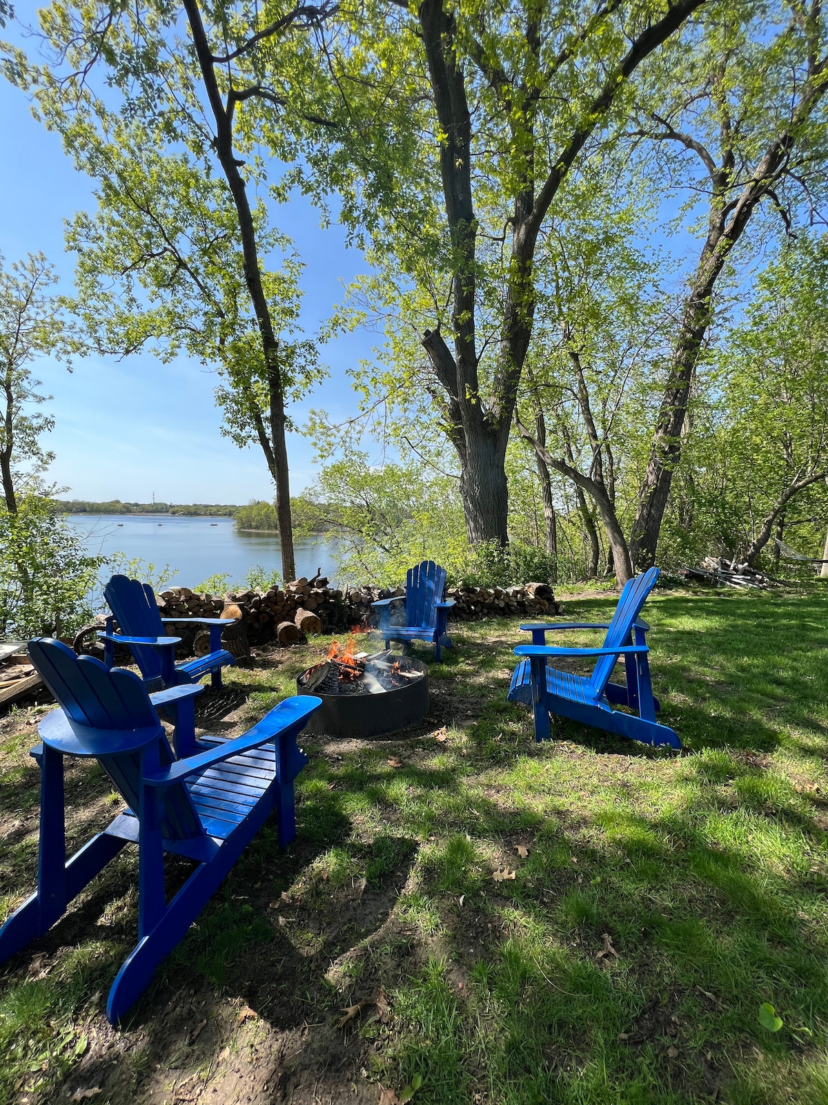 A serene fire pit area is surrounded by four blue adirondack chairs, positioned on grassy ground. Tall trees provide shade, and a stack of firewood is neatly arranged nearby. The tranquil lake is visible in the background, enhancing the outdoor setting.