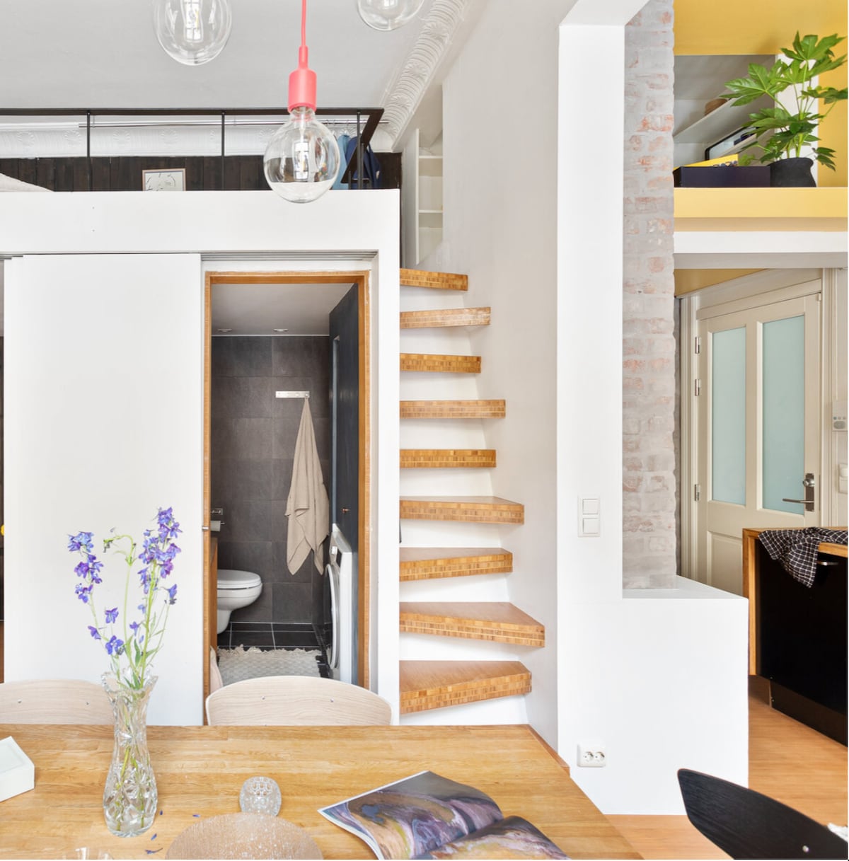 A wooden staircase leads to a lofted area, complemented by a minimalist design. A small vase with purple flowers sits on a wooden dining table, while a bright kitchen area is visible in the background. Natural light enhances the interior's modern appeal.