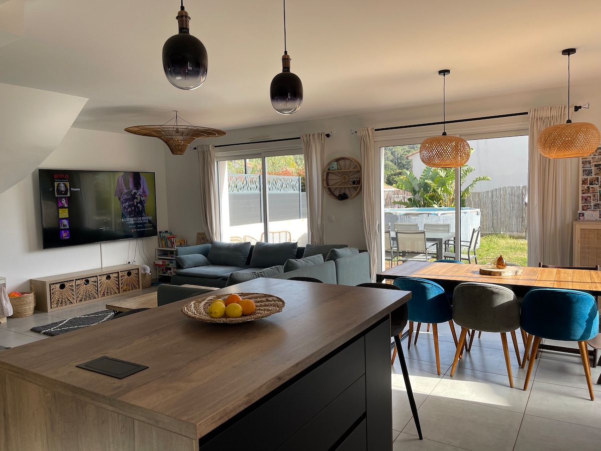 A spacious living area features a large sectional sofa and a wooden dining table surrounded by colorful chairs. Natural light fills the room through sliding glass doors, leading to an outdoor space. Decorative elements include pendant lights and a woven centerpiece on the table.
