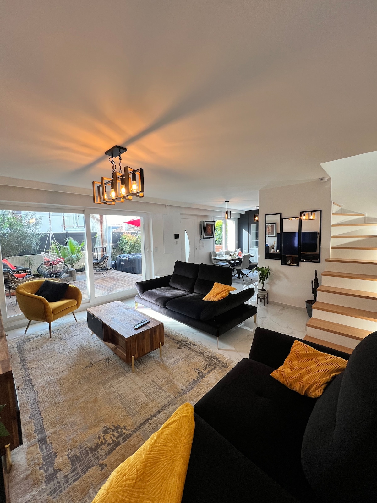 A modern living area features a sleek black sofa adorned with yellow cushions. A wooden coffee table is centered on a patterned rug. Natural light fills the room through large glass doors that open to a terrace, while a stylish chandelier enhances the ambiance.