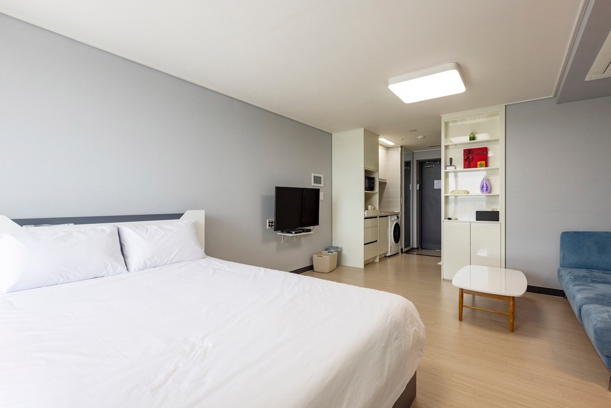 A spacious sleeping area features a large bed with crisp white linens, positioned in front of a wall-mounted television. A small coffee table rests in the foreground, with a compact kitchenette visible in the background, complemented by a cozy blue seating area.