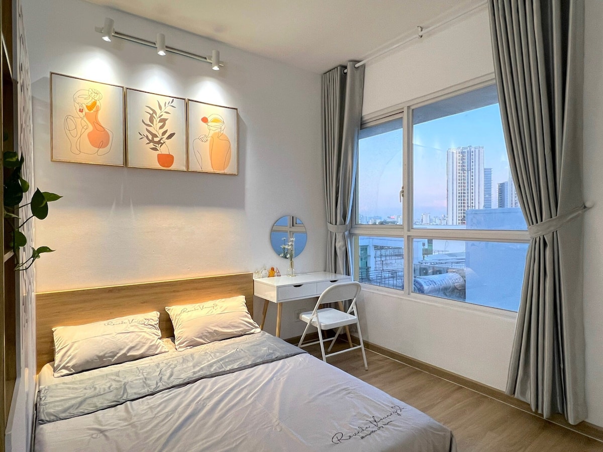 A bright and airy bedroom features a spacious bed with neutral-toned bedding, accented by decorative wall art. A desk with a round mirror and a chair is positioned beside a large window, offering expansive city views and natural light through flowing curtains.