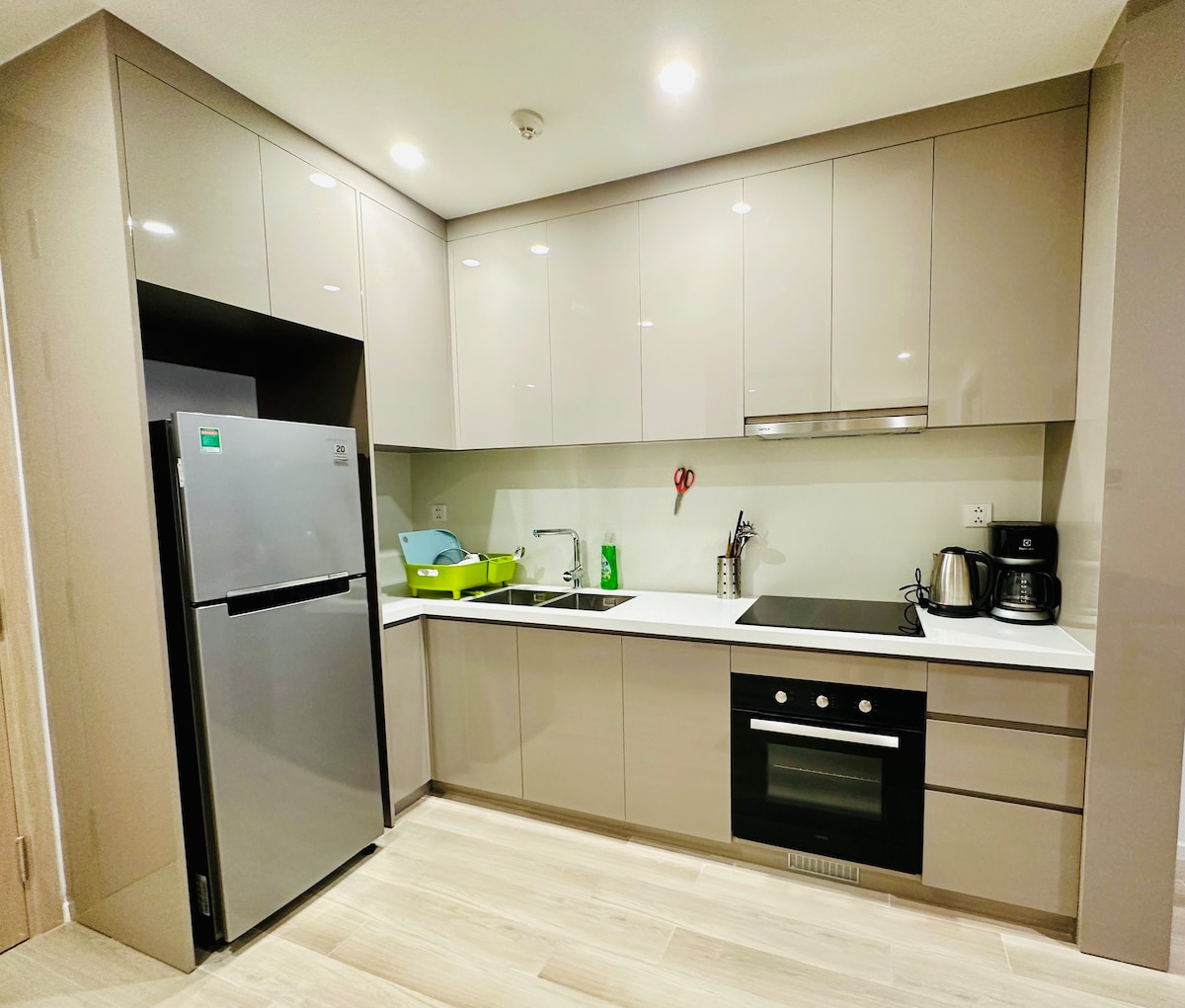 A modern kitchen features sleek gray cabinetry and a white countertop. A stainless steel refrigerator is positioned adjacent to a stovetop and built-in oven. Countertop appliances include a kettle and other kitchen essentials, providing a functional space for meal preparation.