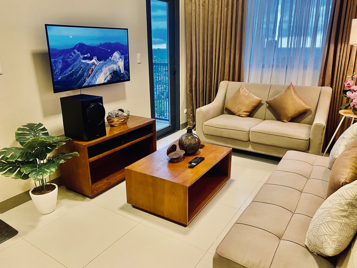 A cozy living area is presented with a soft beige sofa and a matching loveseat, complemented by decorative cushions. A wooden coffee table occupies the center, while a TV is mounted on the wall. Soft natural light filters through sheer curtains, creating a welcoming space.