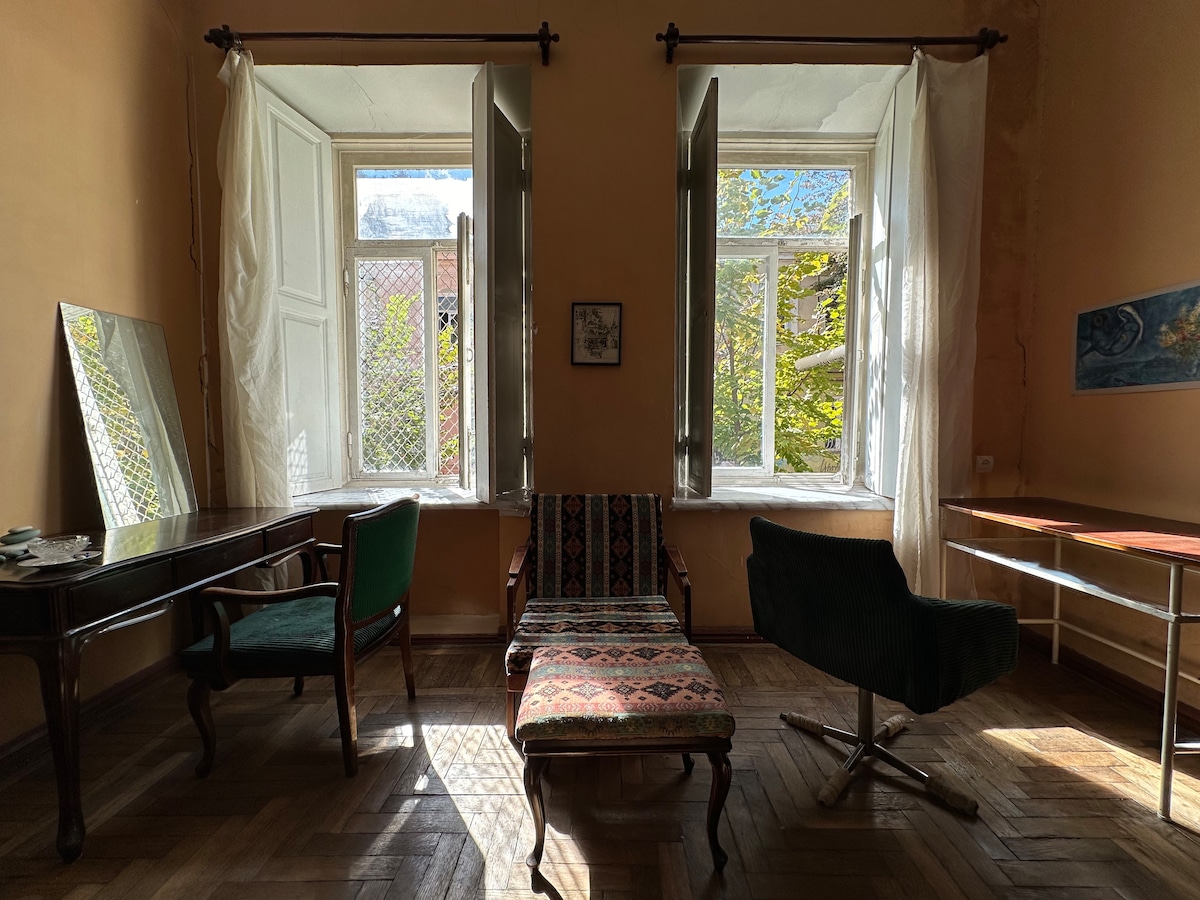 The bright room features two tall windows with flowing white curtains, allowing sunlight to illuminate the space. A vintage chaise lounge is positioned beside a small table, while a mirror and two chairs complete the inviting atmosphere, showcasing the room's unique charm.