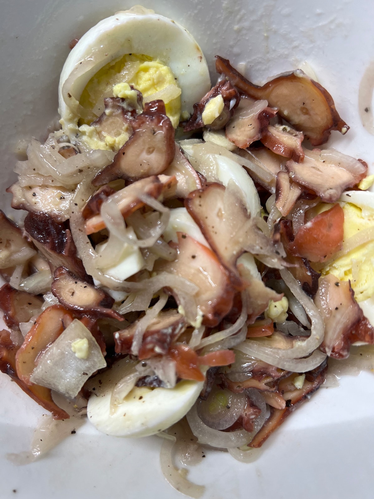 A close-up view of a mixed seafood salad is presented, featuring slices of cooked octopus, chopped onions, and halved boiled eggs. The ingredients are lightly combined, showcasing a balance of colors and textures. A light seasoning is visible, adding to the overall appearance of the dish.