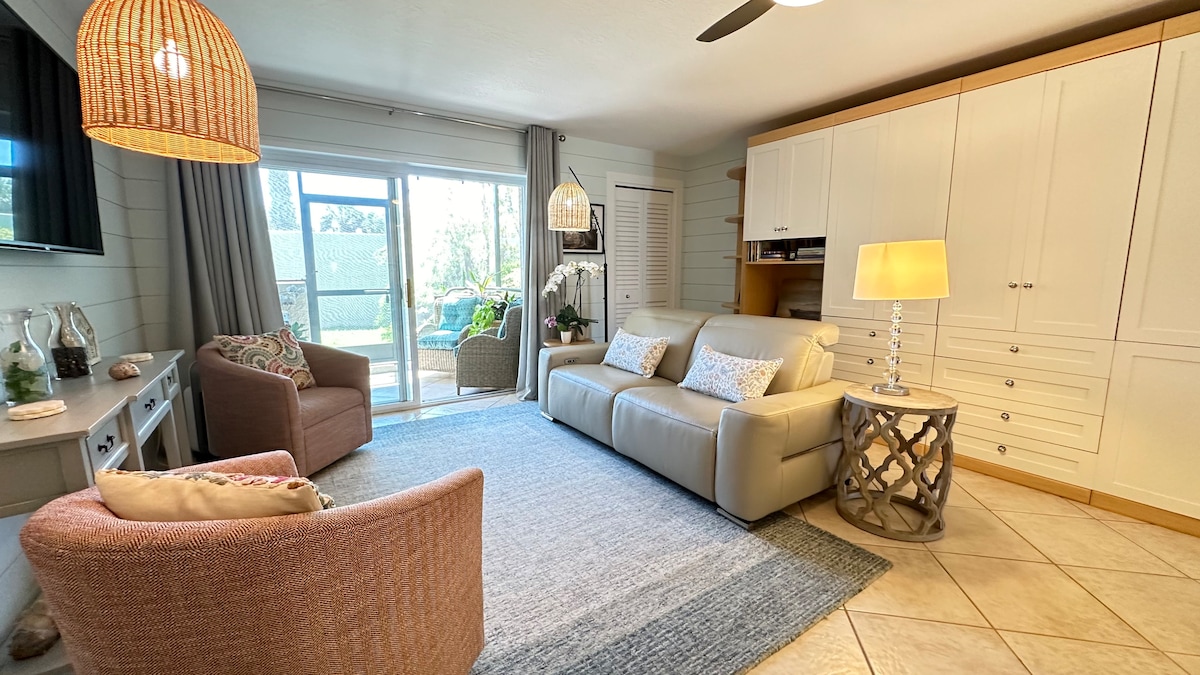The inviting living area features stylish shiplap walls and tile flooring. A comfortable leather electronic reclining sofa is complemented by two swivel chairs. Natural light fills the space through sliding glass doors leading to a screened-in lanai, enhancing the overall ambiance.