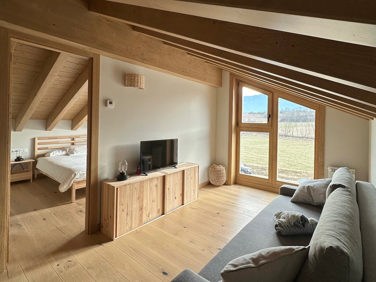 An open-plan loft area features a cozy seating arrangement with soft cushions, complemented by wooden beams and warm-toned flooring. A television is positioned atop a light wooden cabinet. A spacious bed is visible in the adjoining room through a large window, framing scenic outdoor views.