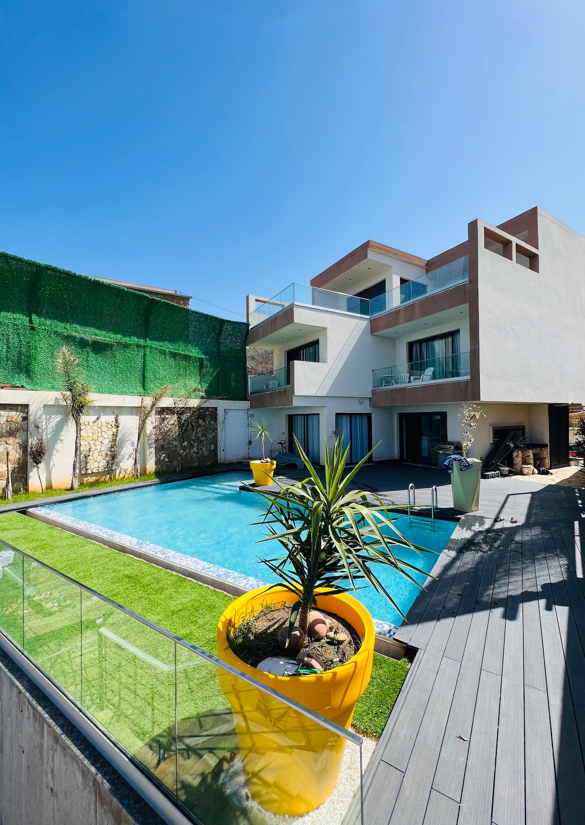The modern villa is showcased against a clear blue sky, featuring swimming pool bordered by lush greenery. A large potted plant adds a splash of color. The architecture features sleek lines and multiple balconies for outdoor relaxation.