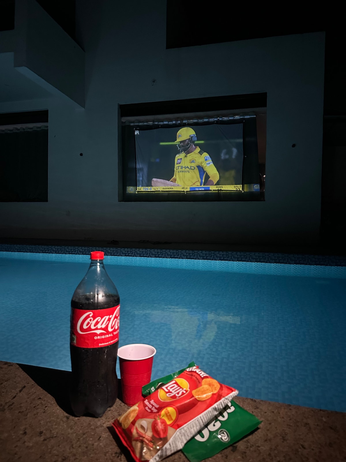 A tranquil pool area is depicted at night, featuring a large screen displaying a sports event. A bottle of Coca-Cola and a red cup are positioned nearby, accompanied by bags of chips on a flat surface, creating a relaxed atmosphere for enjoyment.