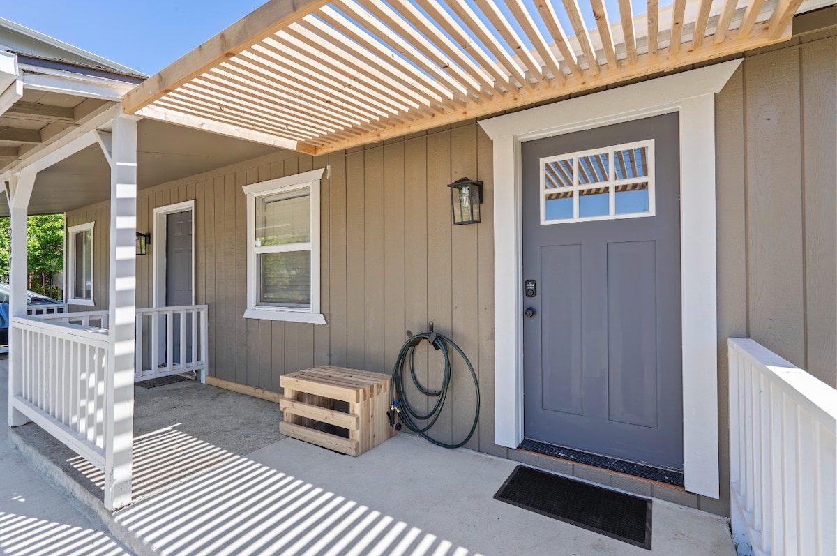 The exterior features a welcoming entrance with a shaded porch and contemporary gray door. Lattice work overhead casts striped shadows on the concrete floor. Nearby, a light fixture is mounted beside the door, and a wooden crate and garden hoses are visible, adding functional elements to the space.