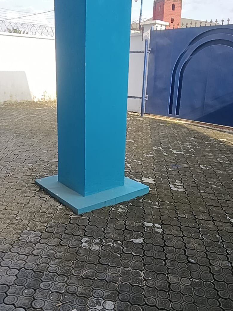 A vibrant blue column stands prominently in an outdoor area with a textured stone pavement. The space is bordered by a white wall and features a solid blue gate in the background, providing a sense of privacy and security.