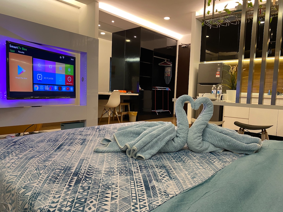 A modern studio space features a spacious bed adorned with a patterned blanket. A smart TV is mounted on the wall opposite the bed, while various amenities are visible in the contemporary kitchen area. Two neatly arranged towels in the shape of swans are placed on the bed.