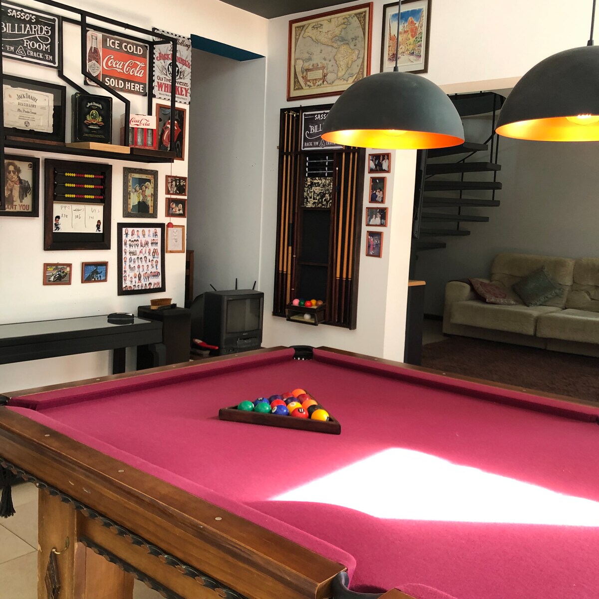 A game room is visible, featuring a vibrant pink pool table with colored balls arranged on it. The walls are adorned with a variety of framed artwork and memorabilia, while soft pendant lights hang above. A cozy seating area is present in the background.
