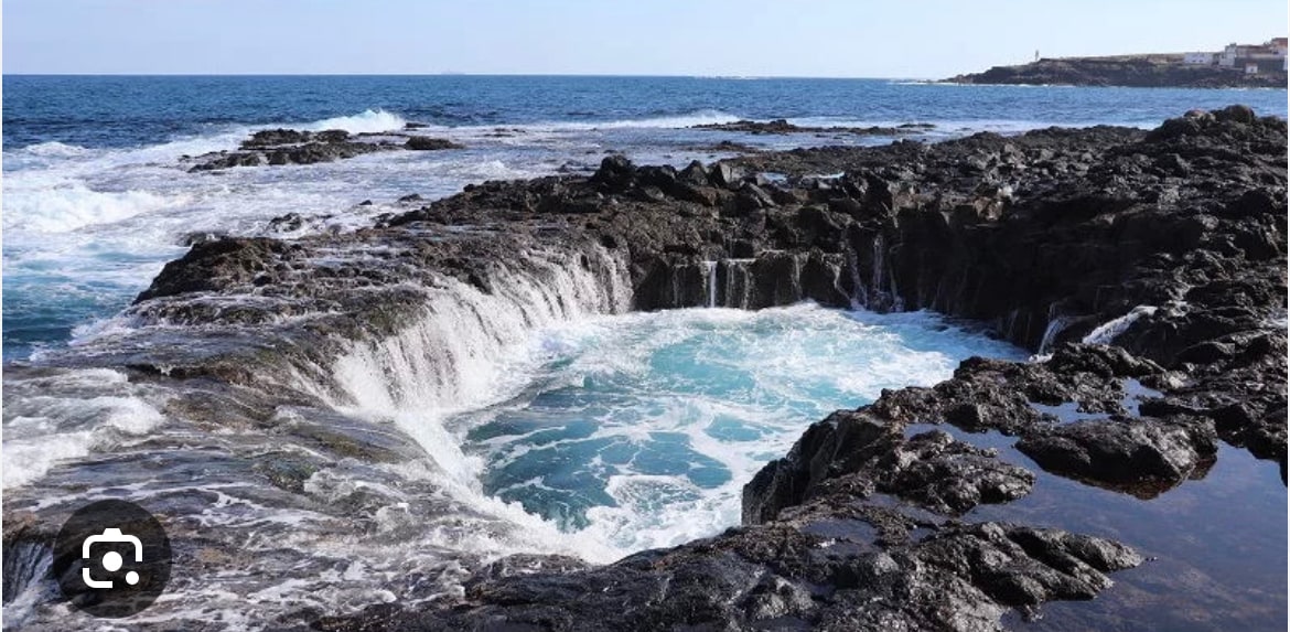 Rugged volcanic rocks surround a natural tidal pool, where waves gently crash against the edges. Clear blue water fills the pool, contrasting vividly with the dark rocks. The horizon features a blue sky with hints of distant coastal landforms.