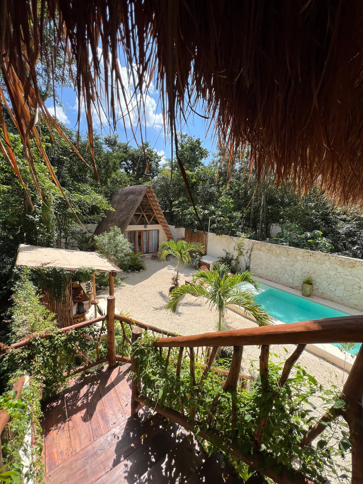 A view of a tranquil outdoor area reveals a sparkling pool surrounded by lush greenery. A charming thatched-roof structure is visible, and palm trees are strategically placed in the pebbled courtyard, creating a serene atmosphere amidst the natural surroundings.