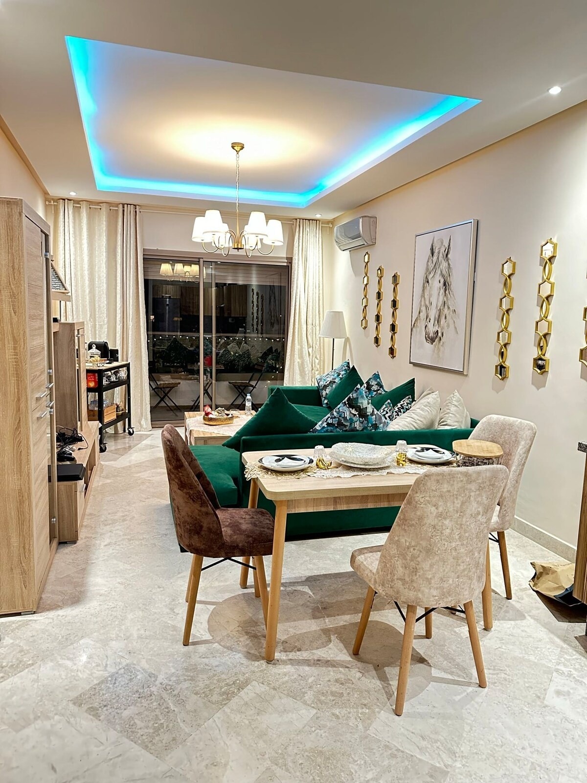 A cozy living area features a green sofa adorned with decorative cushions, positioned opposite a small dining table set for two. Natural light pours in from a balcony, with elegant curtains providing privacy. The ceiling is highlighted by soft blue lighting, complementing the neutral decor.