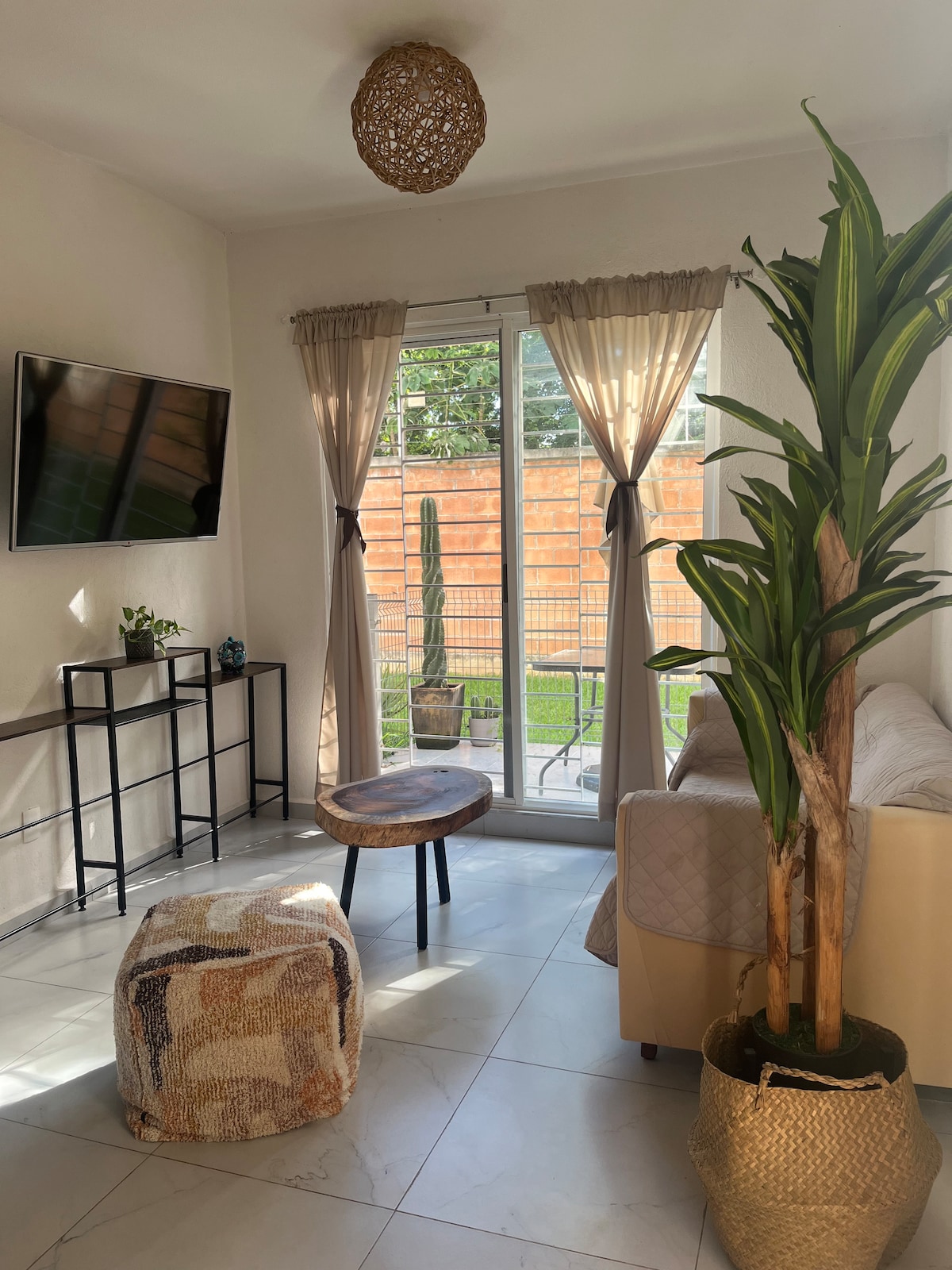 The living area features large glass doors that invite natural light and provide views of the outside terrace. A light-colored couch is complemented by a small round table and a soft ottoman. Decorative plants add a touch of greenery, enhancing the space's inviting feel.