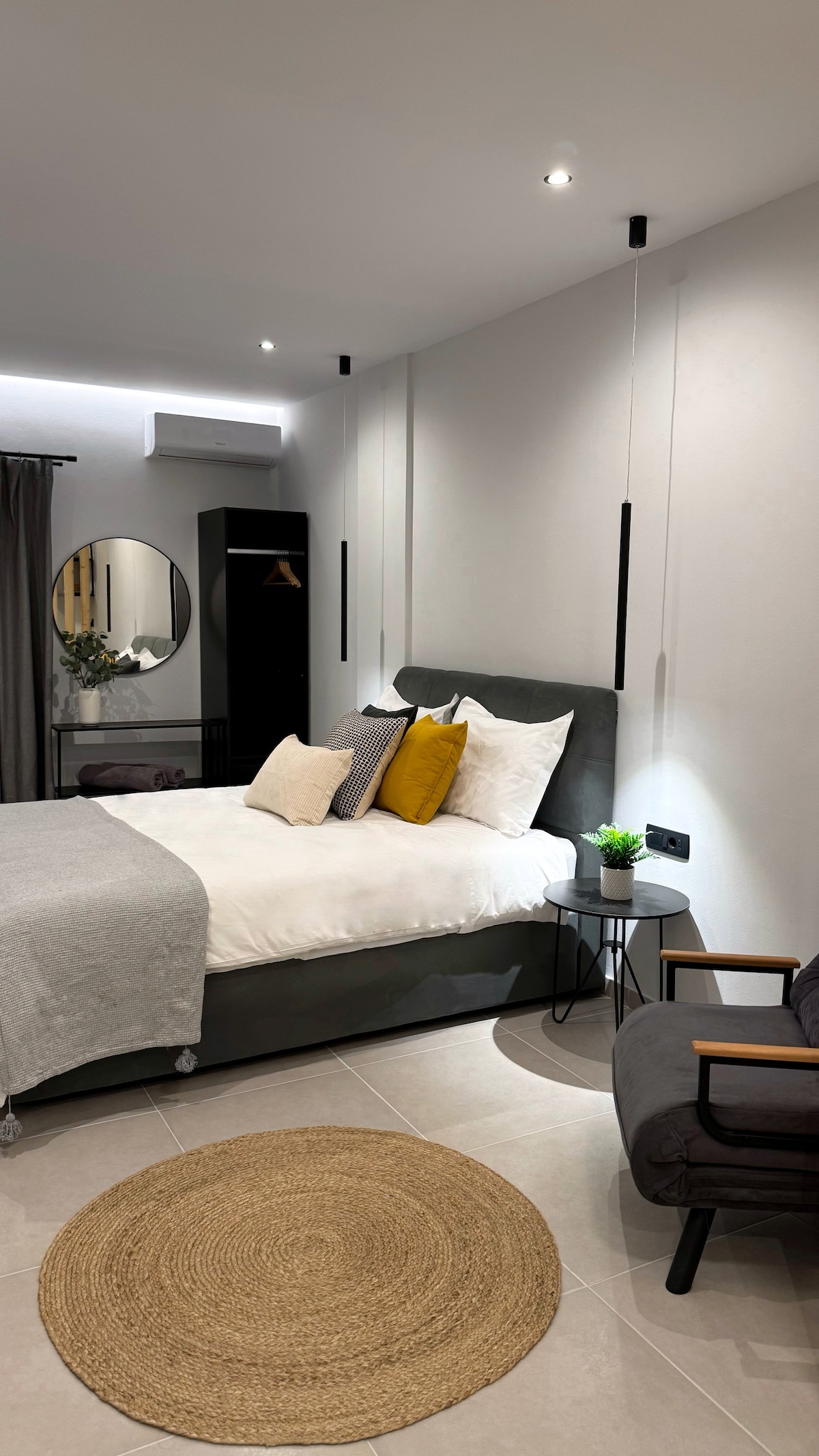 A serene bedroom is featured with a spacious bed dressed in neutral linens and several accent pillows. A round, woven rug complements the room's floor, while a sleek black chair and a mirror add a modern touch. Soft lighting is provided by ceiling fixtures.
