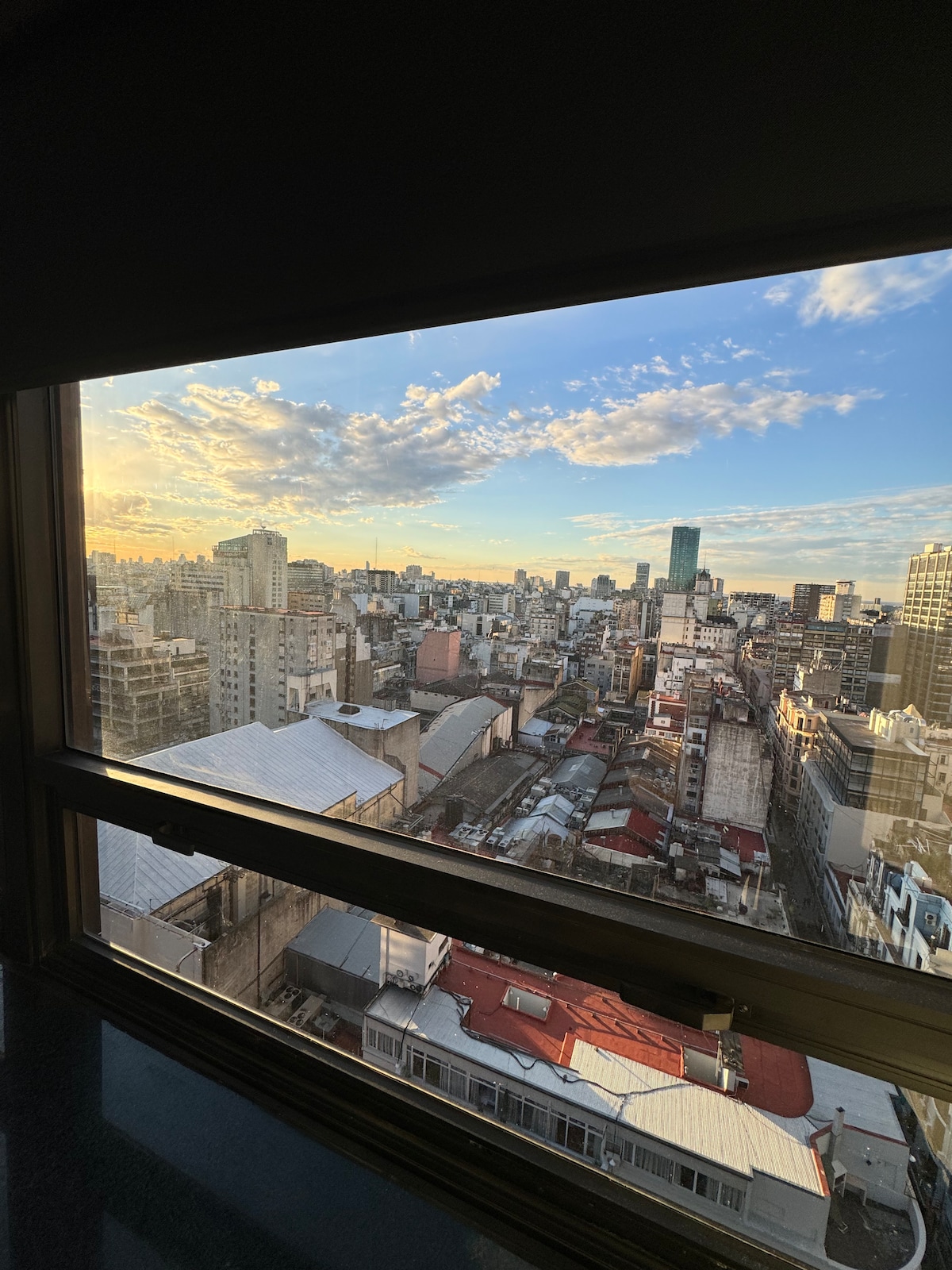 A city skyline is visible through a large window, showcasing a vibrant urban landscape bathed in golden light. The view includes a mix of modern and traditional buildings, with rooftops of varying colors and textures extending across the horizon. Clouds add depth to the sky.