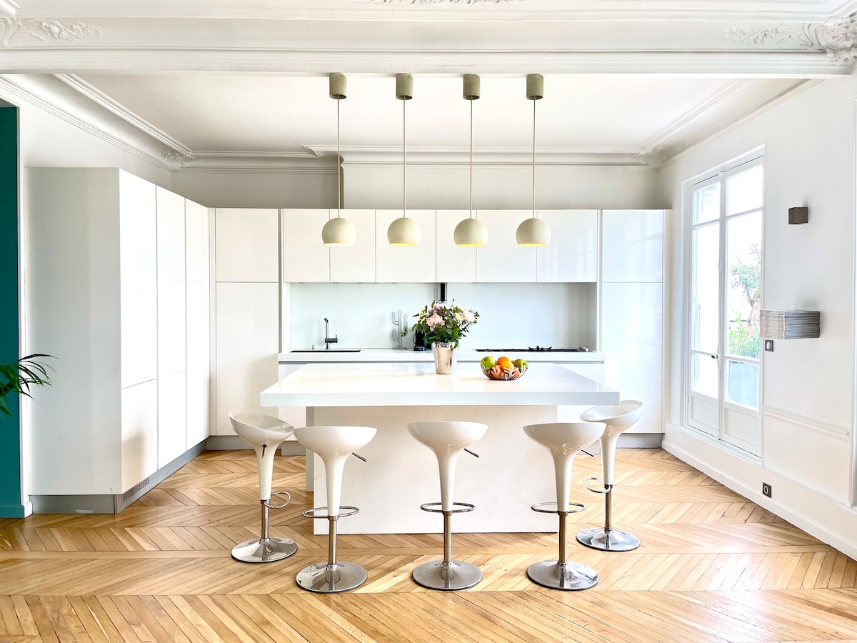 A modern kitchen features white cabinetry and an island with seating for six. Pendant lights hang above the island, illuminating the space. Large windows allow natural light to fill the room, showcasing the herringbone-patterned wood floor.