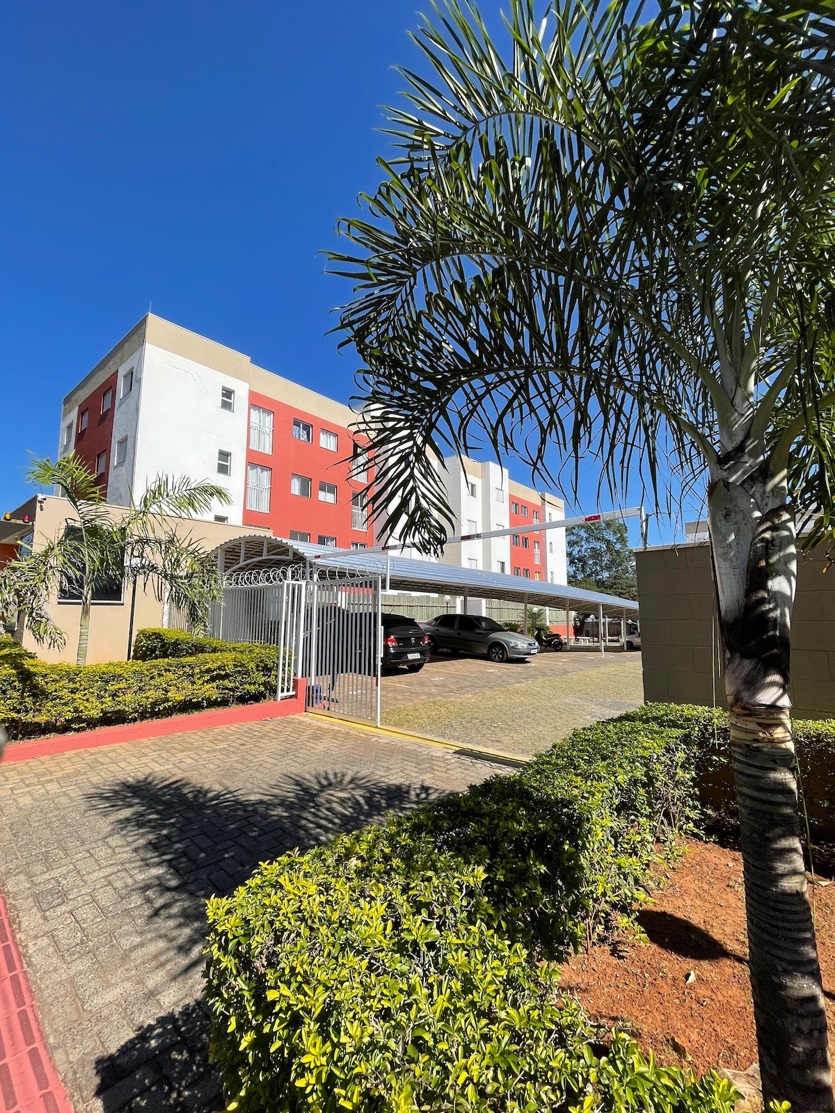 The exterior of a residential building is framed by palm trees and well-maintained shrubs. A gated entrance leads into a paved driveway with shaded parking areas. The building features a mix of red and beige facades under a bright blue sky.