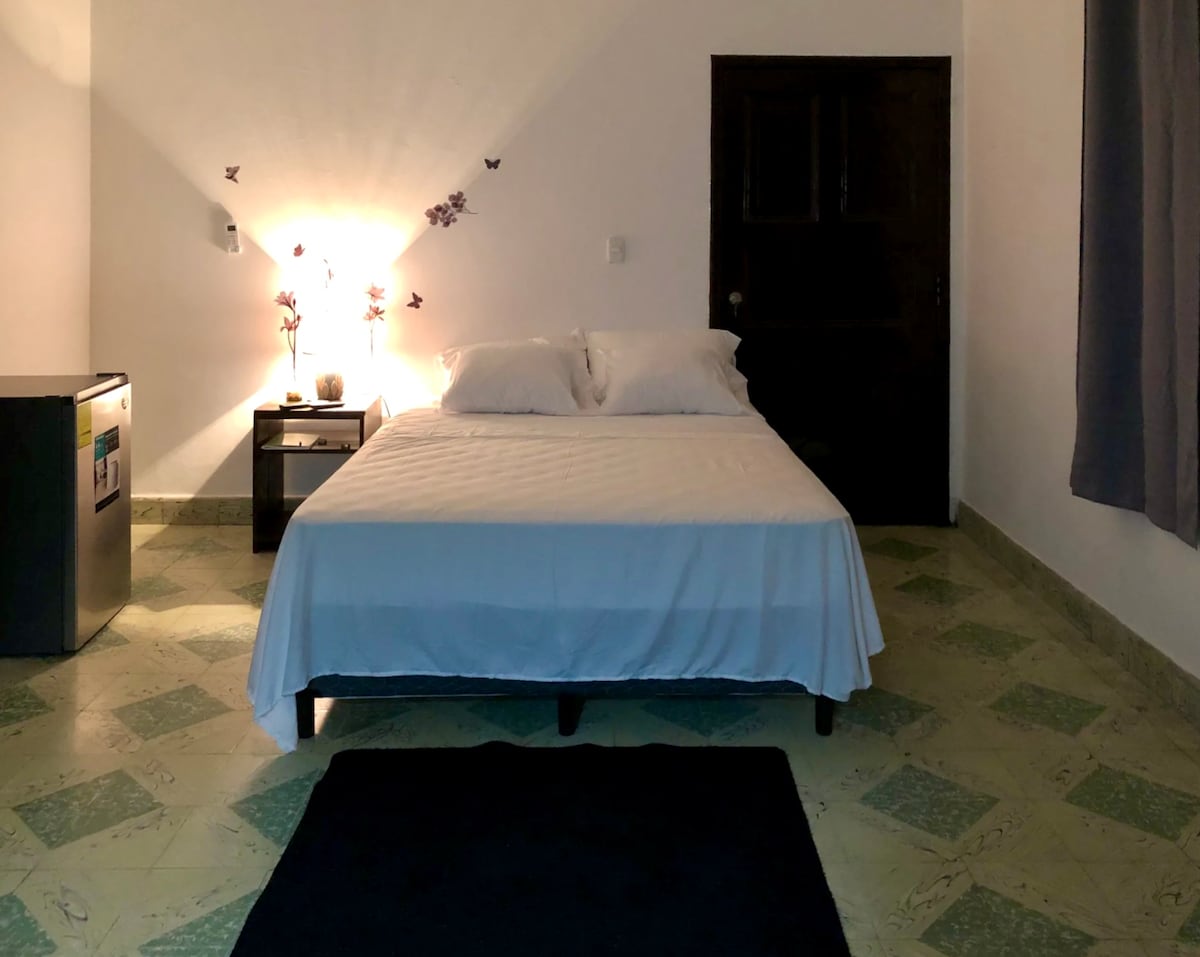 A spacious bedroom features a comfortable bed with white linens, complemented by light from bedside lamps. A small nightstand and a fridge are visible. The textured floor is covered with a dark rug, and simple wall decor adds a touch of warmth to the room.