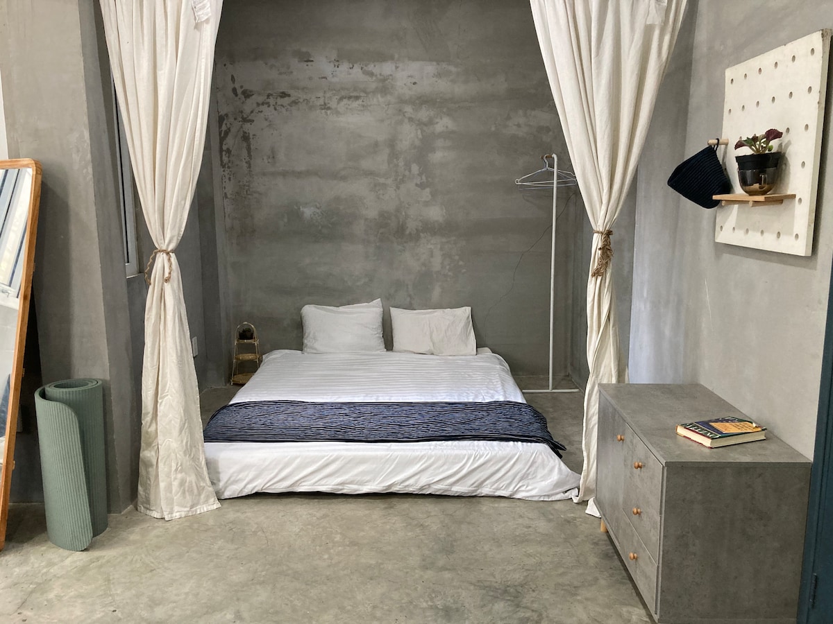 A spacious sleeping area is defined by soft linens and a minimalist aesthetic. The bed is surrounded by light-colored curtains, offering a sense of privacy. A simple storage cabinet and a tall, textured planter are visible, contributing to the uncluttered design.