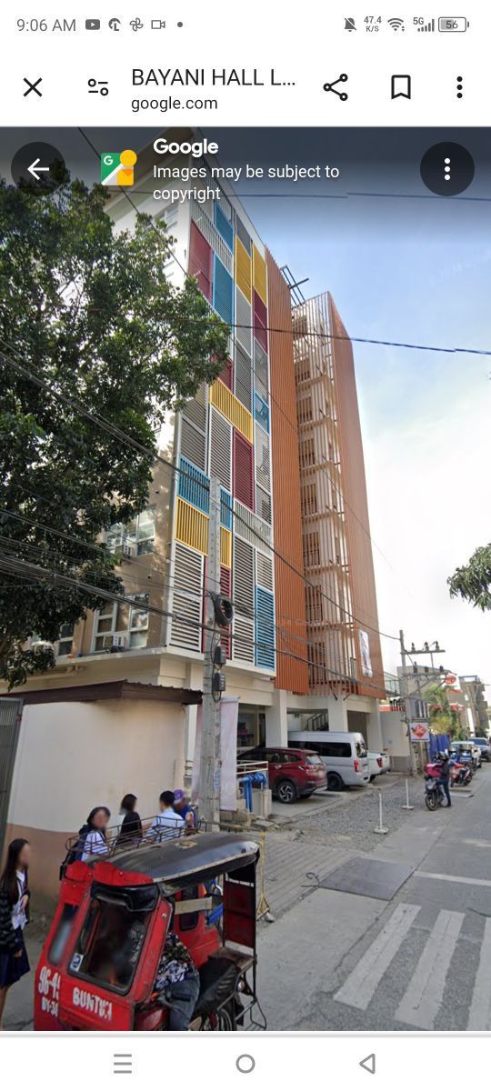 A modern multi-story building is displayed, characterized by colorful panels in shades of blue, yellow, and orange. A covered entrance is visible at the ground level, while nearby vehicles and pedestrians showcase the lively urban environment surrounding the location.