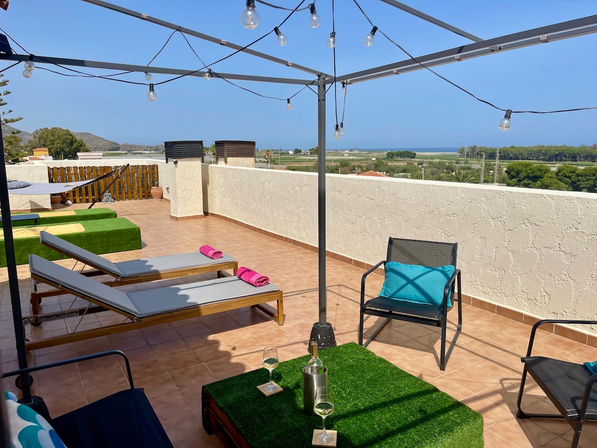 A spacious roof terrace is equipped with outdoor furniture and sun loungers under a canopy. Decorative string lights hang above, creating a relaxed atmosphere. A view of the sea and lush landscape is visible in the background, providing a tranquil setting for relaxation.