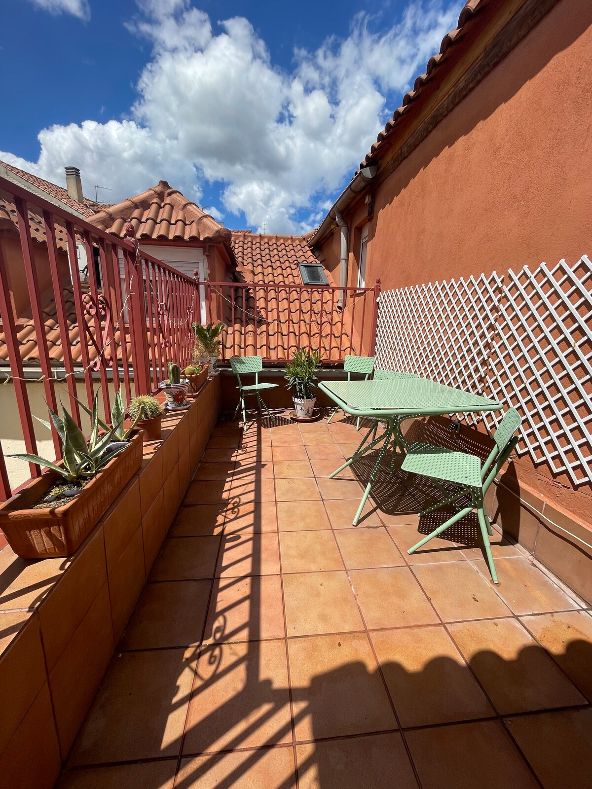 A terrace is shown, featuring green metal furniture arranged on a tiled surface. Potted plants add a touch of greenery, while a lattice panel provides a decorative element. The backdrop includes a red wall and rooftops under a partly cloudy blue sky.