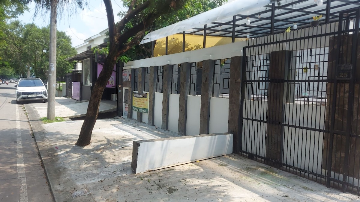 An entrance to a cozy inn is depicted, featuring a secure wrought iron gate with a stone wall surround. A shaded area is provided by a white canopy above the entrance, and a tree is visible along the walkway that leads to the property.