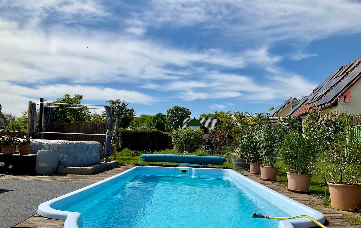 A clear blue swimming pool is surrounded by a wooden deck, with potted plants enhancing the area. In the background, a well-maintained garden is visible, alongside a hot tub and lounge area. The sky features soft clouds, creating a peaceful outdoor setting.