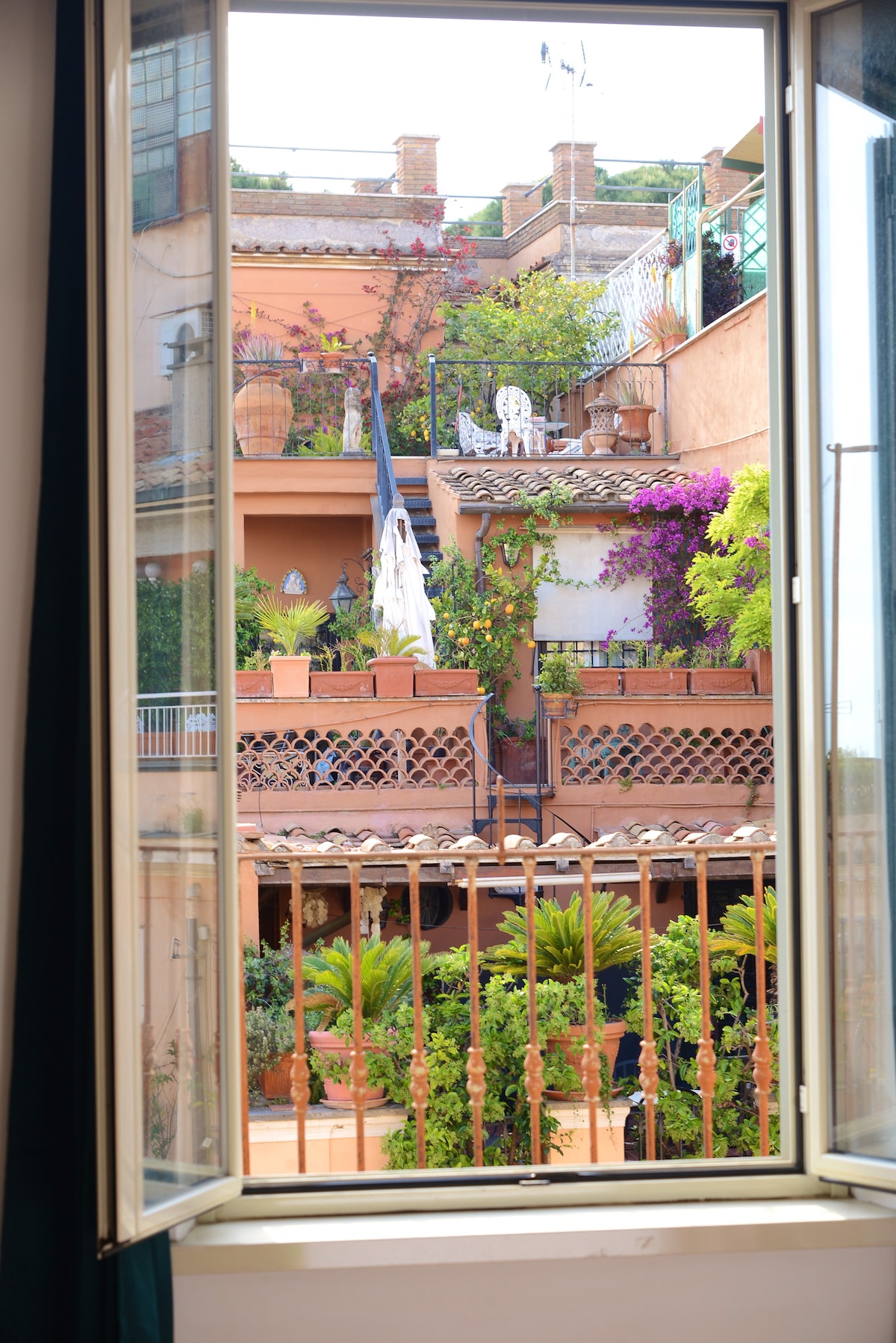 A view from the window showcases a charming terrace adorned with lush greenery and colorful plants. The vibrant bougainvillea climbs the walls, while elements of terracotta decor blend with the natural surroundings. A glimpse of outdoor seating is visible, creating a serene outdoor ambiance.