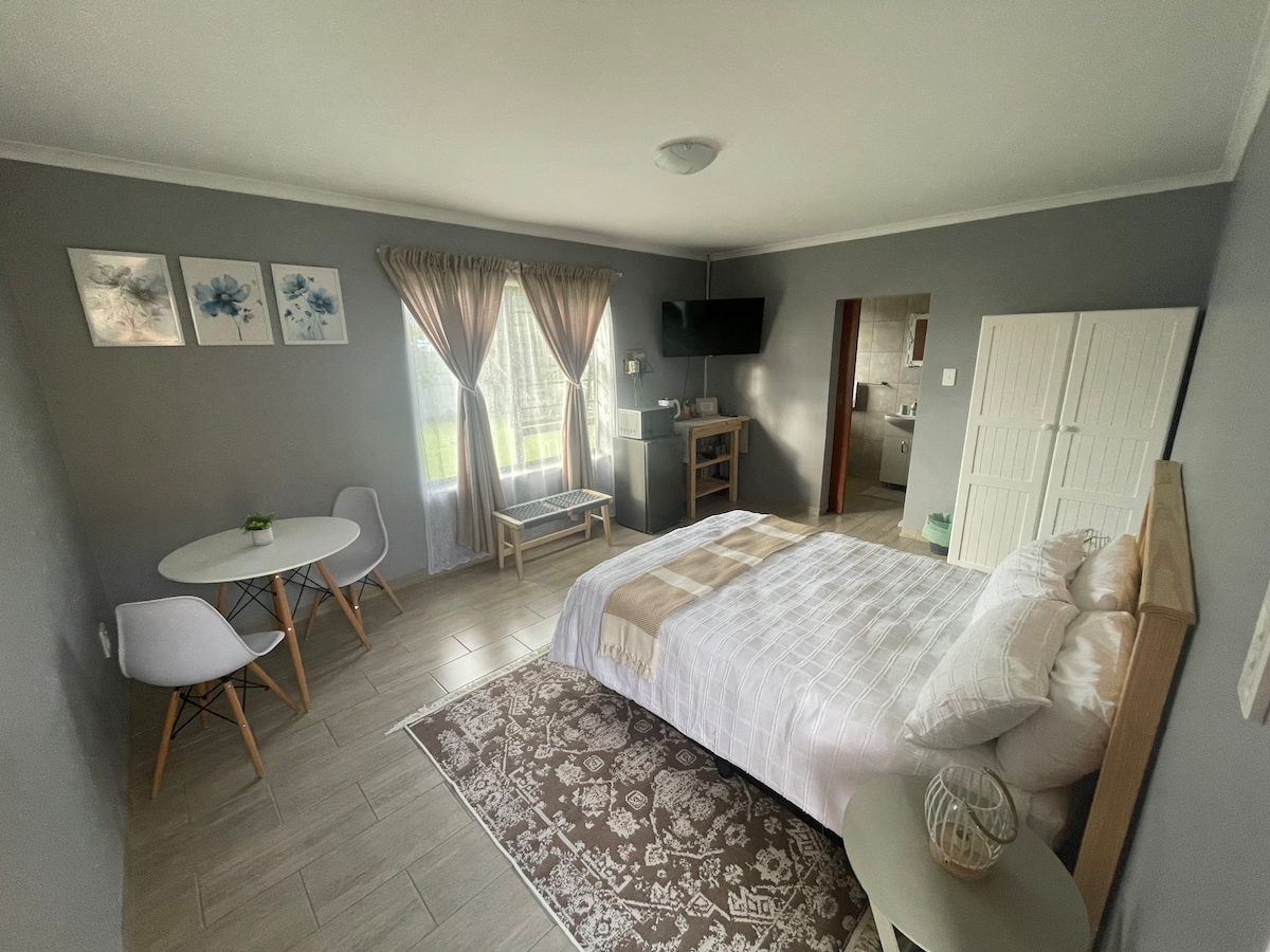 A cozy studio apartment features a spacious bed with neutral bedding, situated next to large windows draped with soft curtains. A small dining area with two chairs and a table is present, along with a TV mounted on the wall and a doorway leading to the en-suite bathroom.