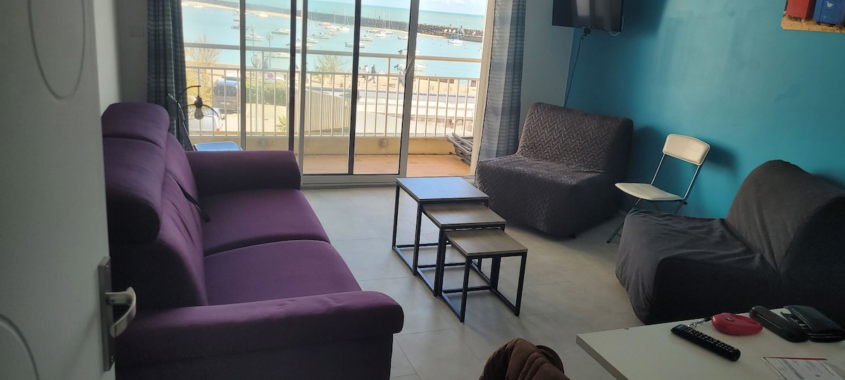 A bright living area is showcased with a purple sofa and two casual seating options. A set of three coffee tables are arranged in front. Large glass doors allow views of the sea and connect to a balcony, enhancing the space's openness.