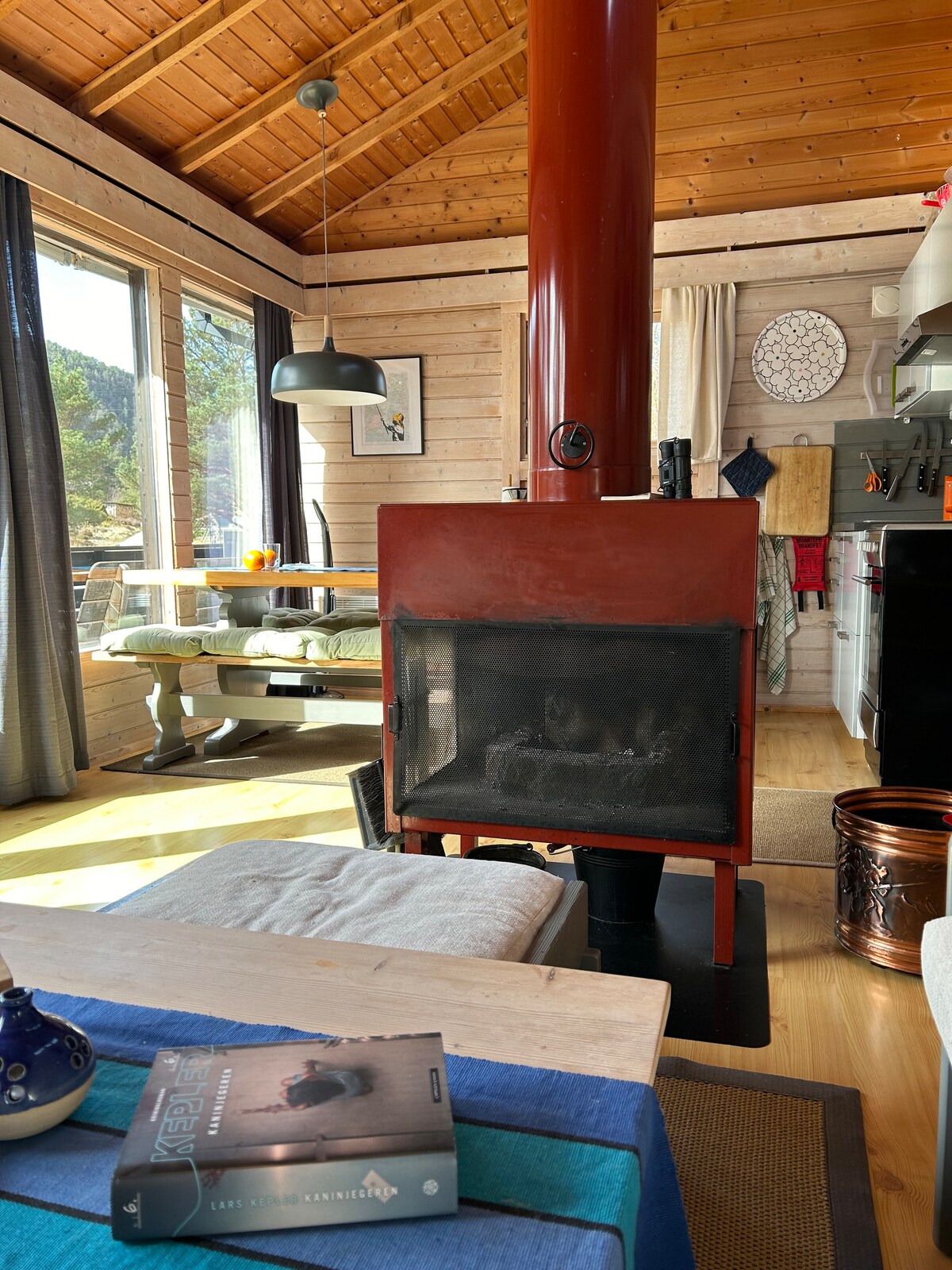 A cozy living area features a red wood stove as its focal point, surrounded by large windows that invite natural light. A dining table is visible, complemented by a green seating area. Soft furnishings and decor create a comfortable and welcoming environment.