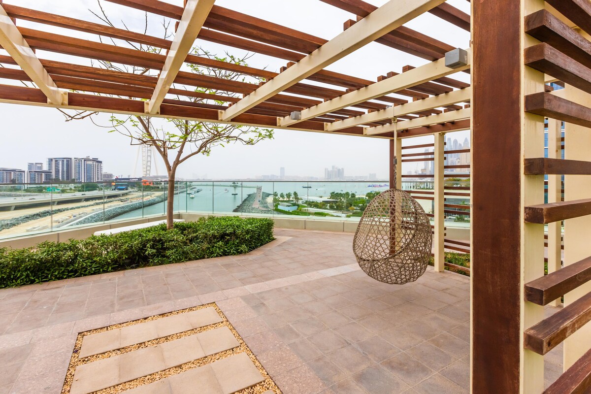 An outdoor terrace with a wood and metal pergola offers views of the water and skyline. A woven hanging chair is positioned on a stone-paved surface. Lush landscaping borders the terrace, enhancing the outdoor space's natural feel.