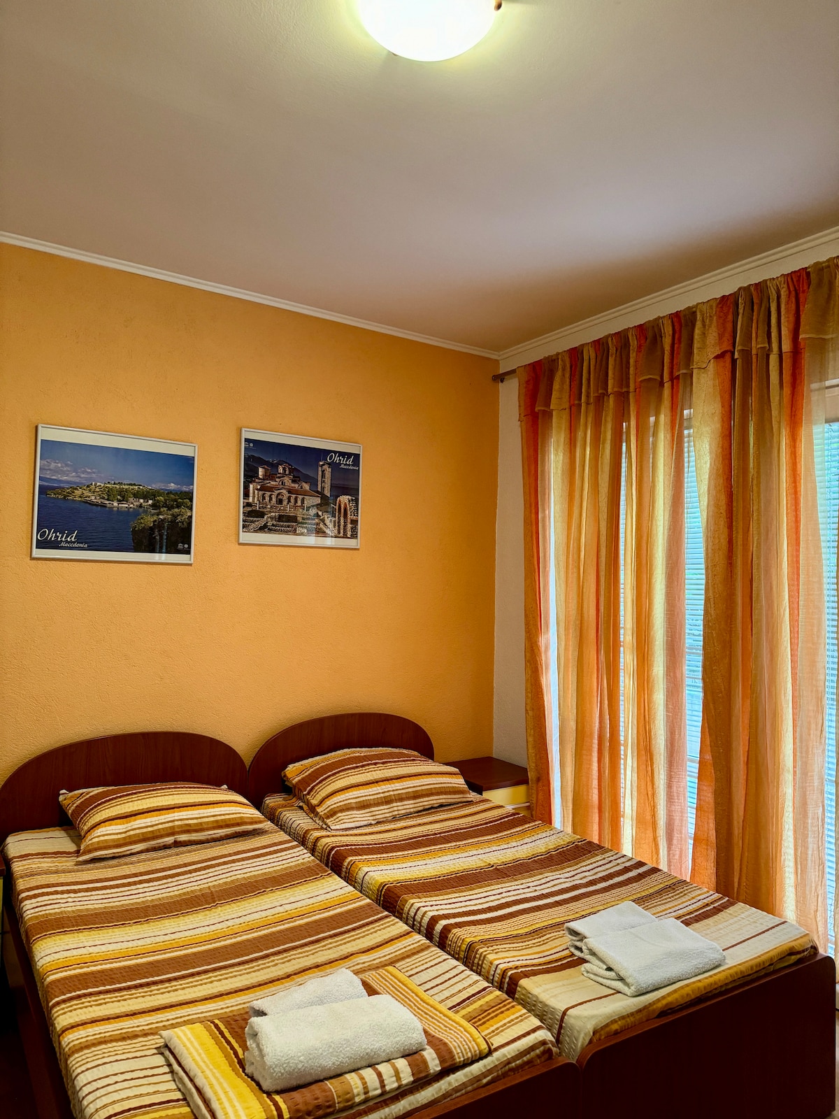 A well-lit bedroom features two twin beds, each adorned with striped bedding. Light brown walls complement the warm orange curtains, which partially filter natural light. Framed pictures hang above the beds, adding a decorative touch to the space. Fresh towels are neatly placed on the beds.