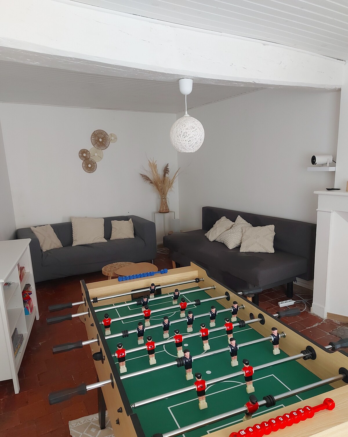A cozy living area is highlighted by a foosball table at the center. Two gray sofas with cushions are positioned against the wall, complemented by a coffee table and decorative wall elements. Natural light enhances the inviting atmosphere of the space.