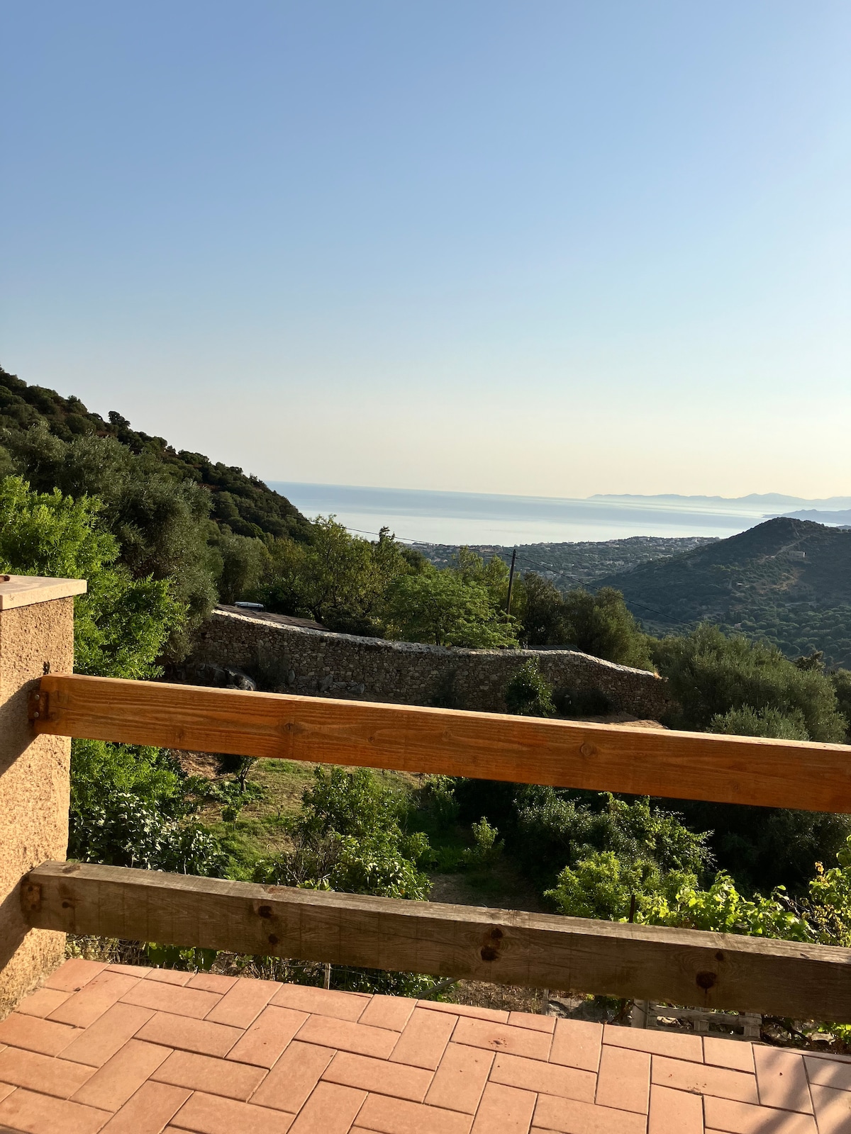A scenic view is presented from the terrace, showcasing lush greenery and rolling hills leading to the coastline. The distant sea glistens under the sunlight, while a sturdy wooden railing frames the view. The landscape embodies a serene and tranquil atmosphere.