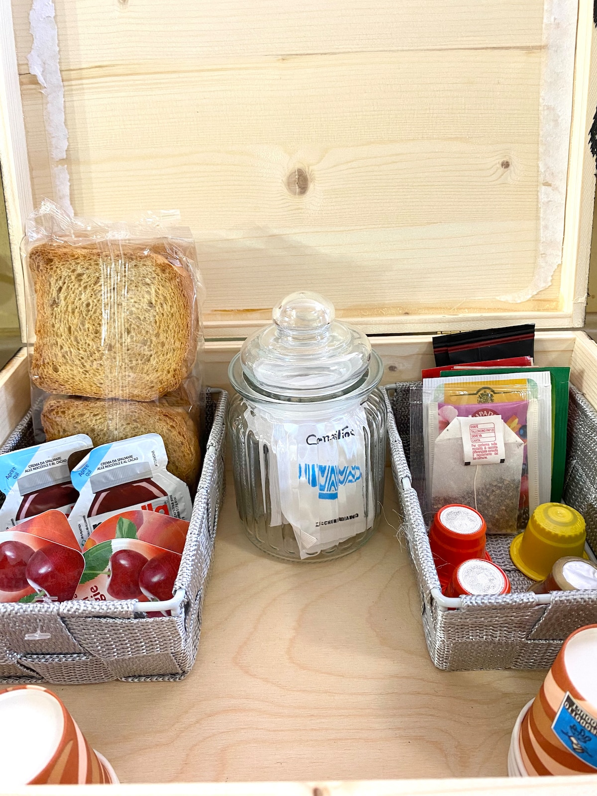 A wooden storage box is open, displaying organized snacks and beverages. Two woven containers hold various spreads and sweet toppings. A glass jar contains tea bags and coffee capsules, while diverse packets and small condiment containers fill the space. The setting conveys a thoughtful hospitality.