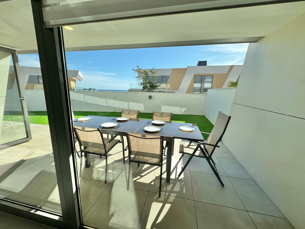 An outdoor dining area is presented, featuring a spacious table set with plates and six chairs. A backdrop of lush green grass and clear skies enhances the tranquil setting. Large sliding doors connect this area to the interior space, allowing for seamless transition.