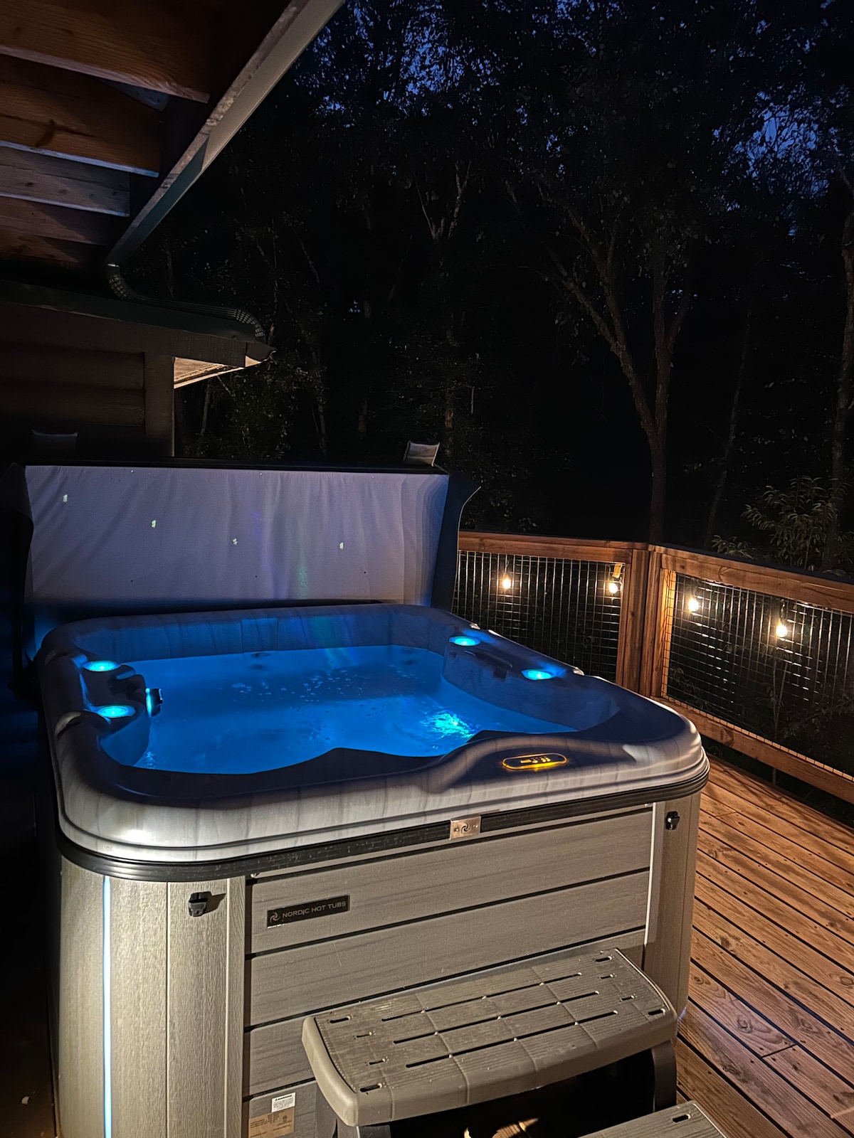 A hot tub is illuminated by soft lighting on a wooden deck, creating a tranquil setting surrounded by trees. The tub is filled with water and features jets, providing a perfect space for relaxation under the night sky.