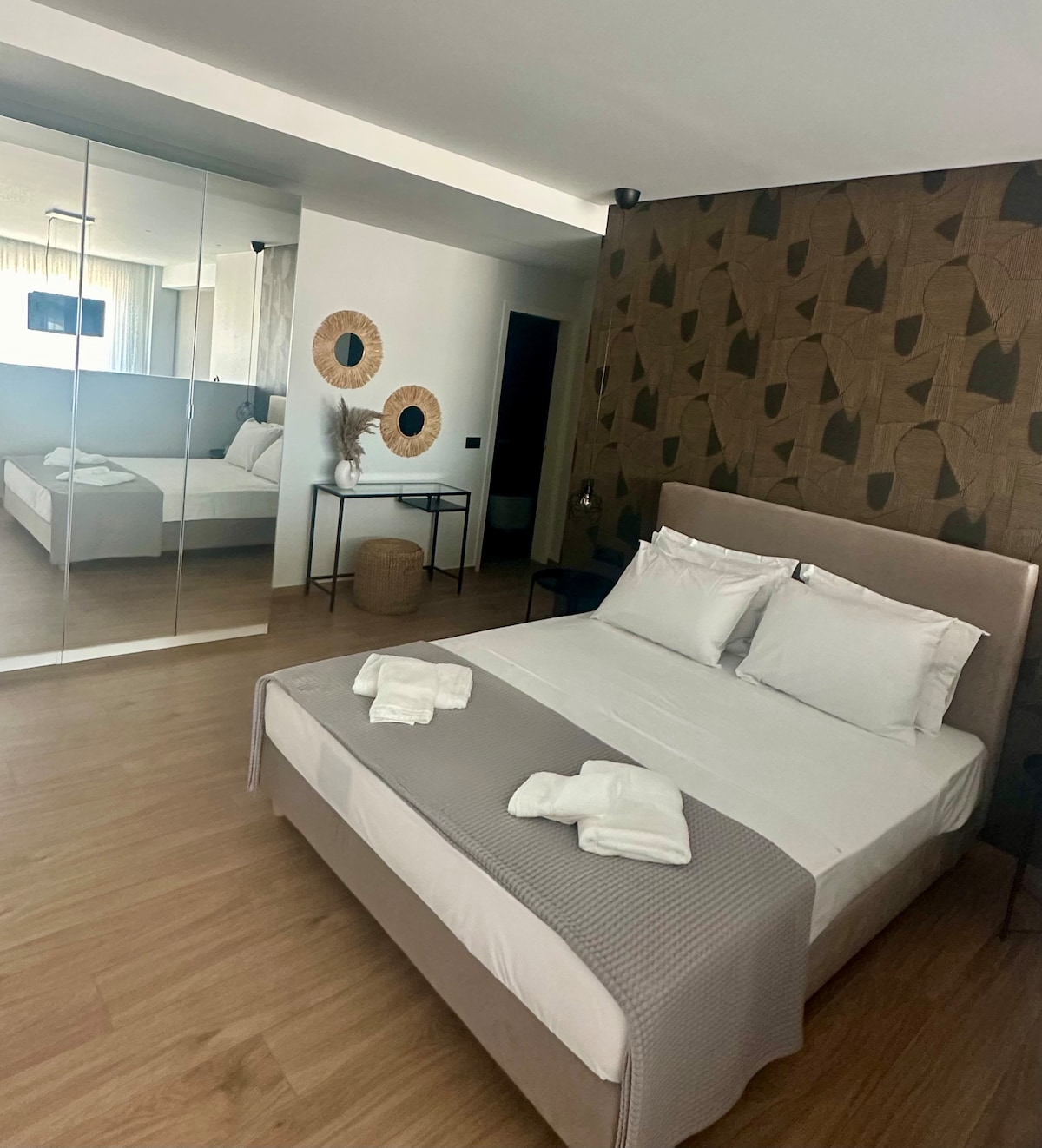 A modern sleeping area is equipped with a spacious bed dressed in neutral linens. Surrounding wall decor includes circular woven pieces. A mirrored wardrobe enhances the room's openness, and light enters through a window draped in sheer curtains.