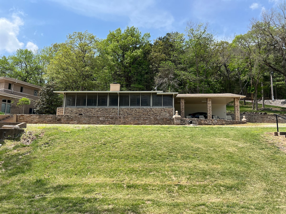 The exterior view of the remodeled lakefront property showcases a stone façade and expansive screened porch. Lush green grass leads to a gentle slope with trees in the background. A covered parking area is visible to the side, providing additional shelter.