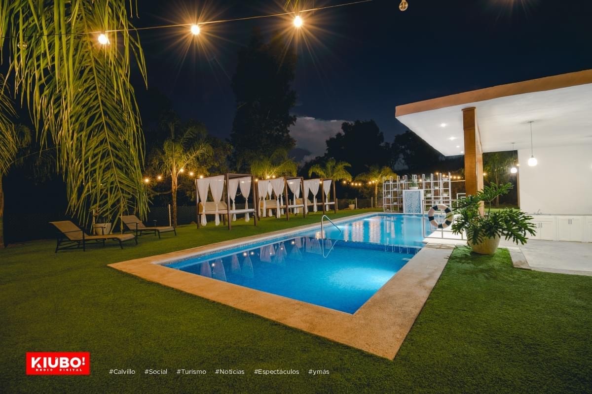 An outdoor pool area is illuminated by string lights, enhancing a relaxing atmosphere. A clear, rectangular pool is surrounded by lush greenery, with comfortable lounge chairs positioned beside it. White cabanas provide shade nearby, complementing the tranquil landscape under the evening sky.